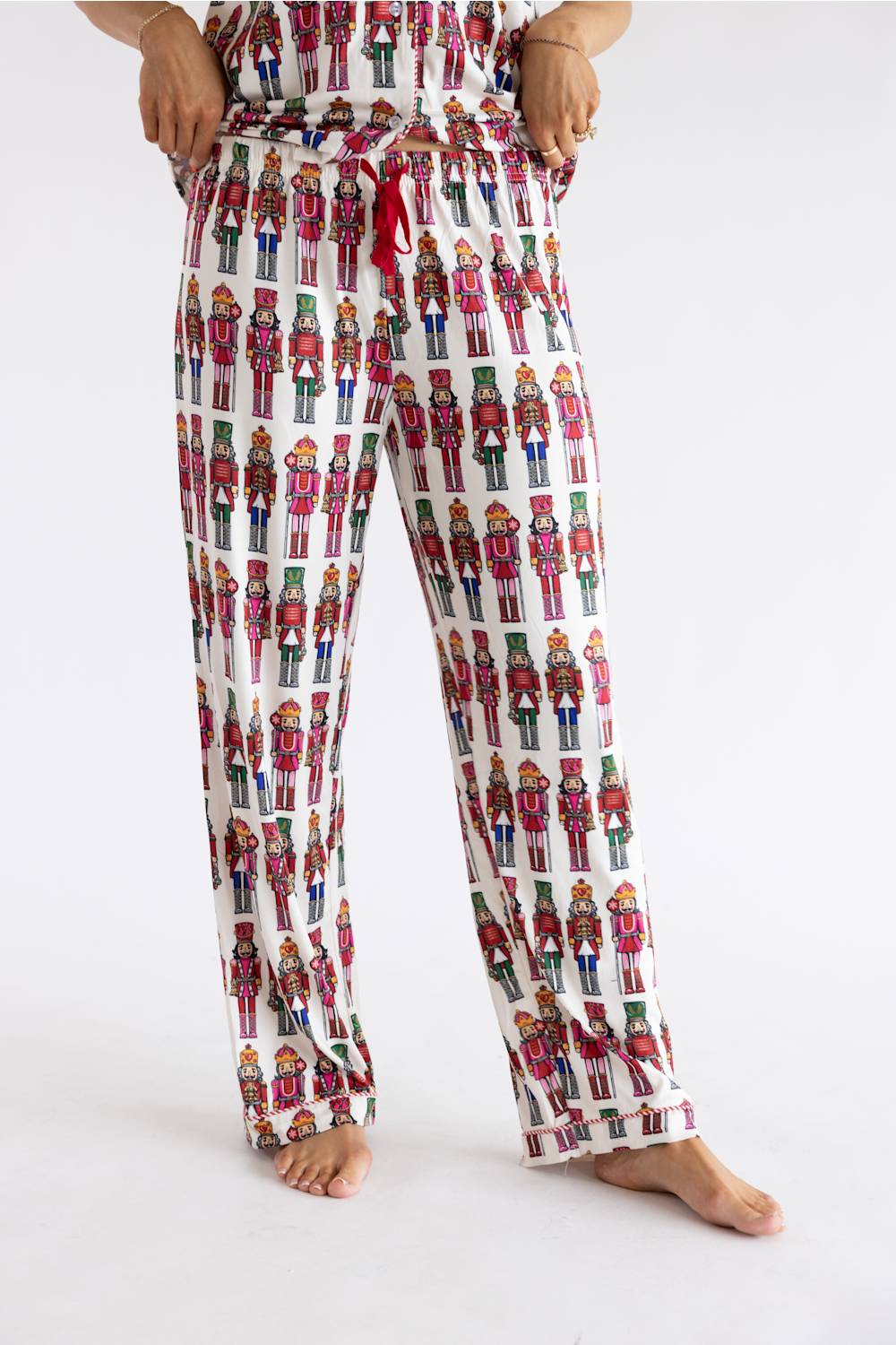 A person wearing white pajama pants covered in a repeating nutcracker soldier print, shown from the waist down against a plain white studio background.