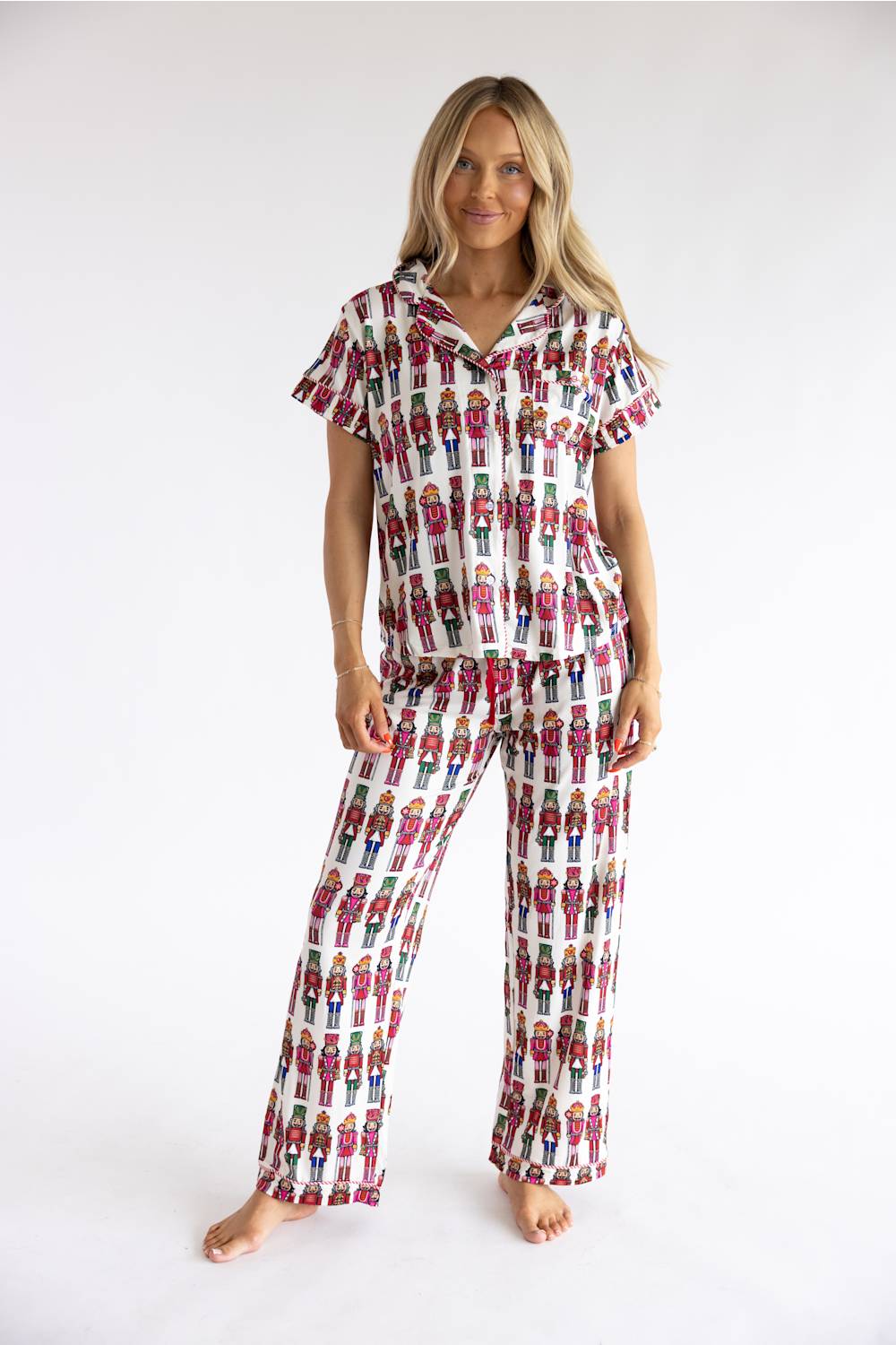 A woman modeling a matching short-sleeve button-up pajama top and wide-leg pajama pants set, both featuring an all-over repeating print of colorful cartoon soldiers on a white background, against a plain white studio backdrop.