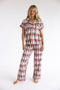 A woman modeling a matching short-sleeve button-up pajama top and wide-leg pajama pants set, both featuring an all-over repeating print of colorful cartoon soldiers on a white background, against a plain white studio backdrop.