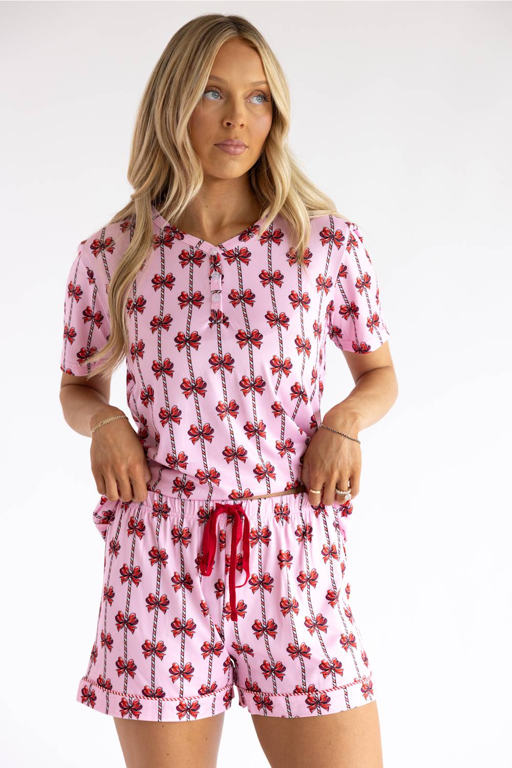 A woman modeling a pink short-sleeve button-front top and matching shorts set with a repeating red bow print, photographed against a plain light background.