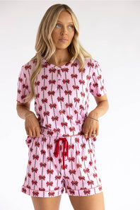 A woman modeling a pink short-sleeve button-front top and matching shorts set with a repeating red bow print, photographed against a plain light background.