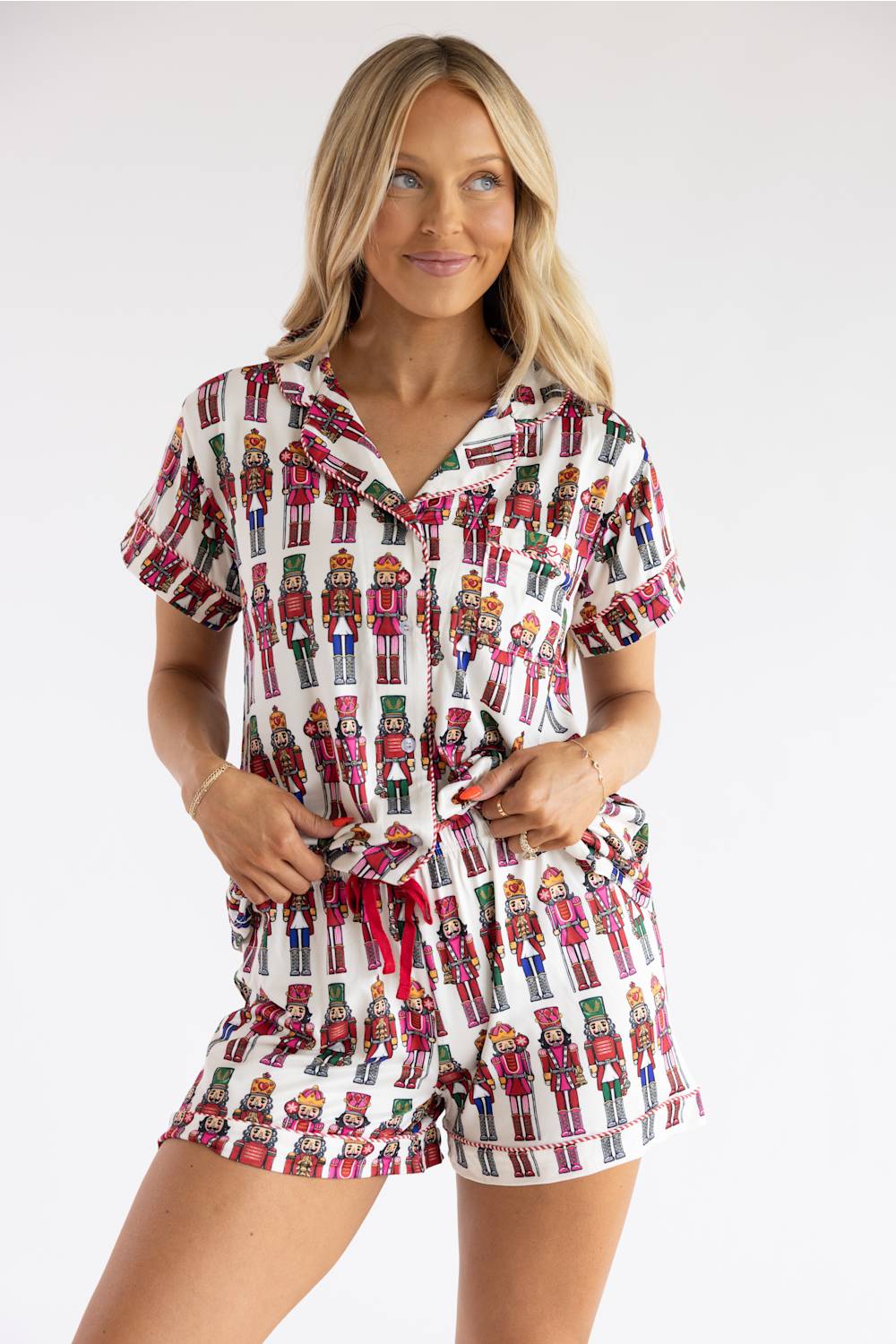 A woman modeling a matching short-sleeve button-up shirt and shorts pajama set with an all-over nutcracker print on a white background.
