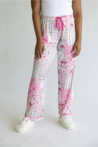 A person wearing a pair of pajama pants with a pink and white patchwork print, featuring a pink drawstring waist and white slippers, shown against a plain light gray background.