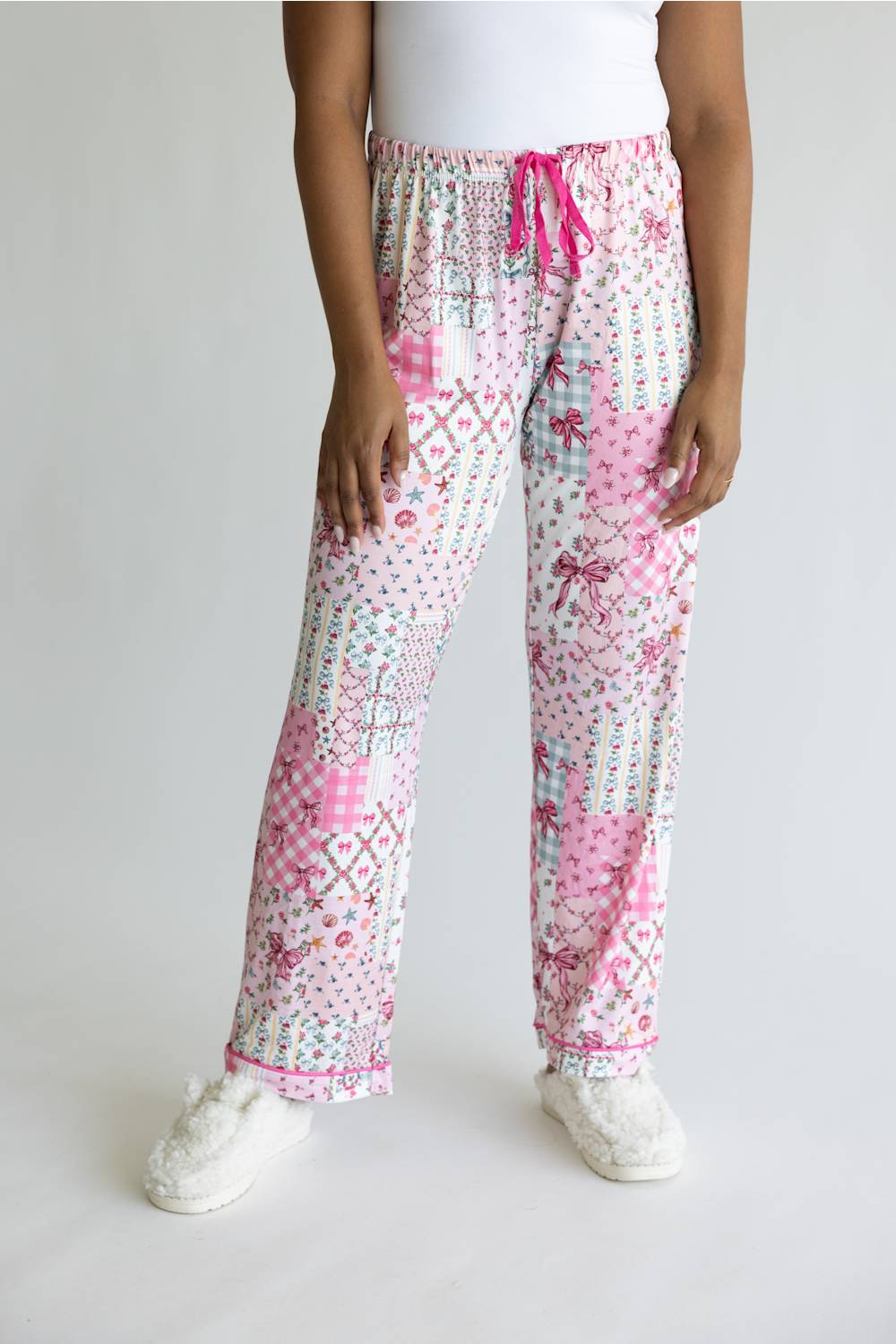 A person wearing a pair of pajama pants with a pink and white patchwork print, featuring a pink drawstring waist and white slippers, shown against a plain light gray background.