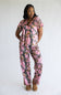 A woman modeling a pink camouflage print short-sleeve pajama set with a matching top and pants, paired with brown suede slippers, against a plain white studio background.