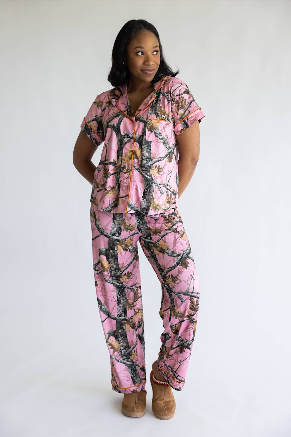 A woman modeling a pink camouflage print short-sleeve pajama set with a matching top and pants, paired with brown suede slippers, against a plain white studio background.