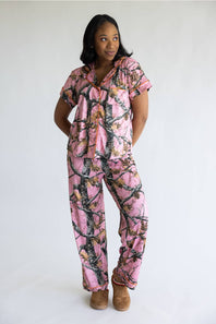 A woman modeling a pink camouflage print short-sleeve pajama set with a matching top and pants, paired with brown suede slippers, against a plain white studio background.