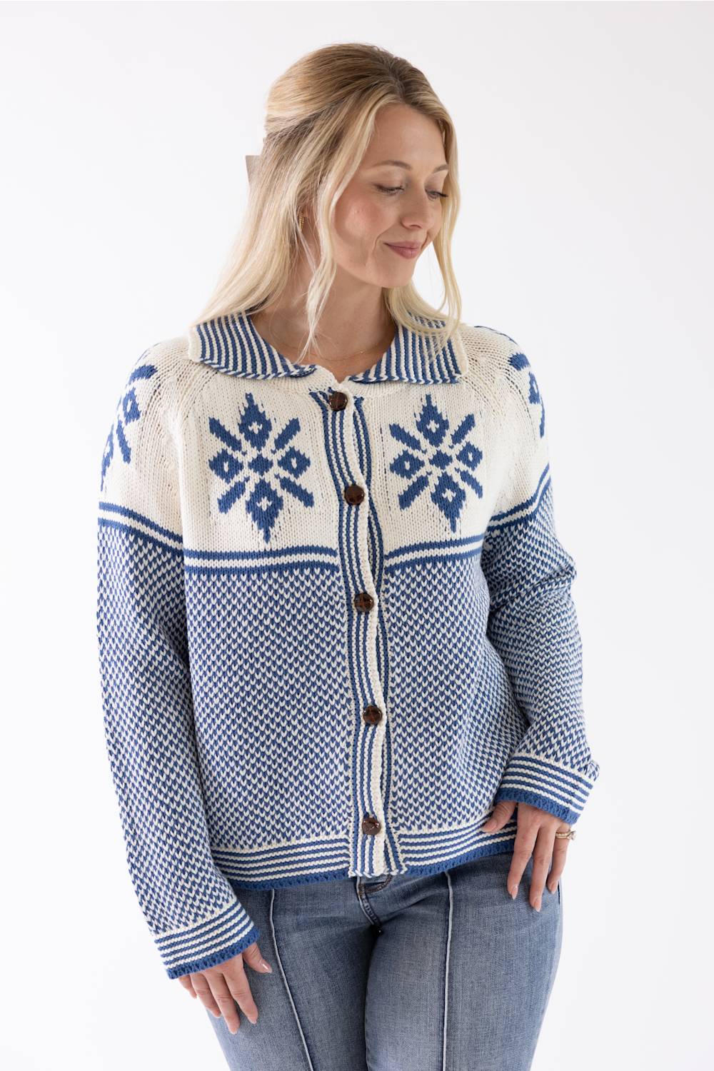 A woman wearing a blue and cream knit cardigan sweater with a button-front closure and Nordic snowflake patterns, paired with blue jeans against a plain white background.