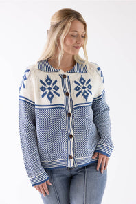 A woman wearing a blue and cream knit cardigan sweater with a button-front closure and Nordic snowflake patterns, paired with blue jeans against a plain white background.