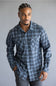 A man wearing a long-sleeve button-up plaid shirt in shades of blue, gray, and white, paired with gray pants, standing against a plain gray studio background.