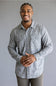 A smiling man wearing a light gray and white plaid long-sleeve button-down shirt, standing against a solid gray studio background.