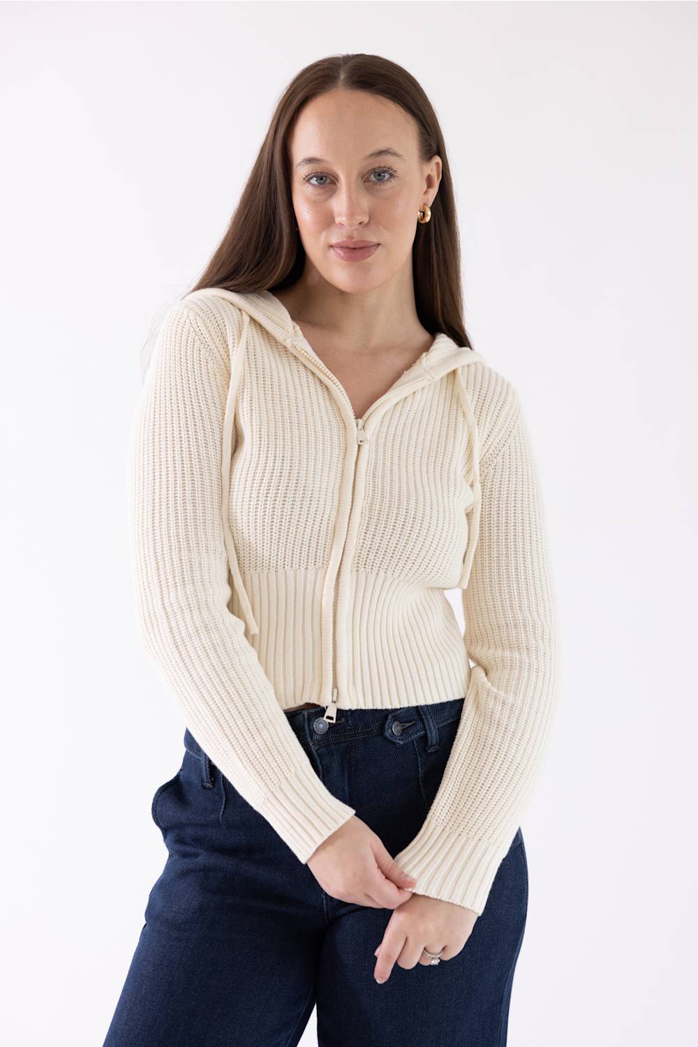 A woman modeling a cream ribbed knit zip-up hooded sweater with front pockets, paired with dark blue jeans, against a plain white studio background.