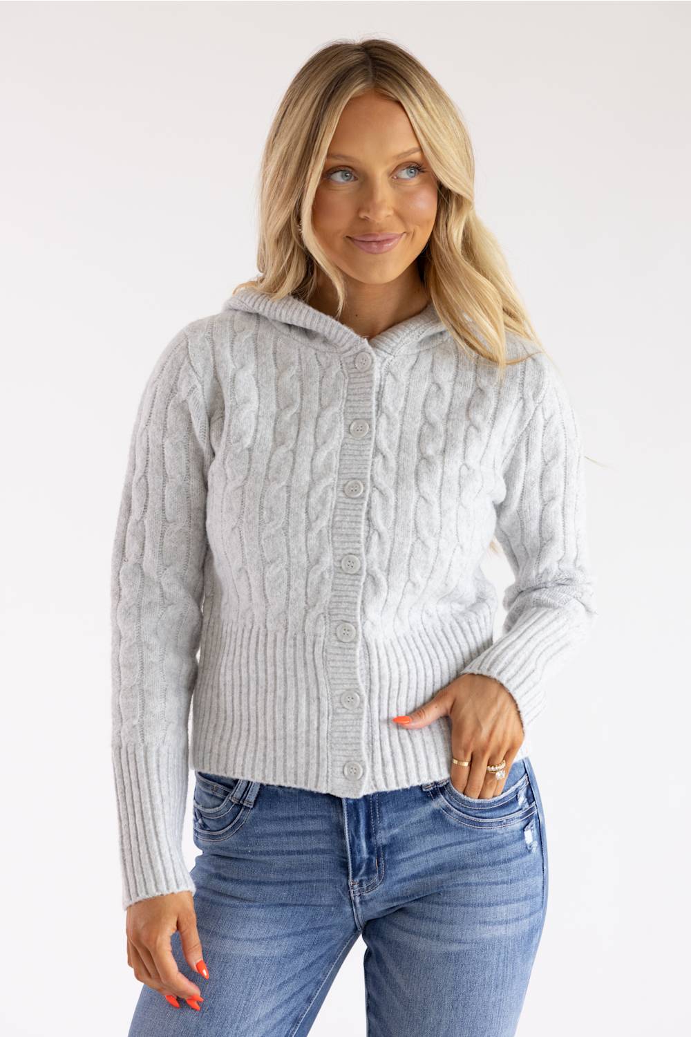 A woman wearing a light gray cable-knit hooded cardigan sweater with blue jeans, standing against a plain white studio background.