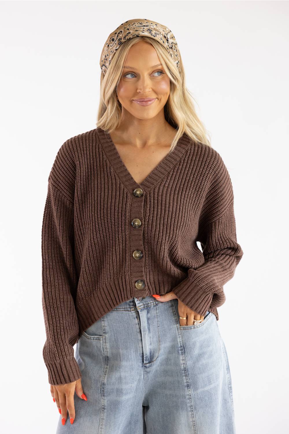 Front view of a smiling woman with wavy blonde hair wearing a printed headband, a dark brown V-neck button-down cardigan sweater, and light-wash jeans.
