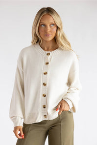 A blonde woman wearing a cream-colored knit cardigan sweater with brown buttons, paired with olive green pants, against a plain light gray background.
