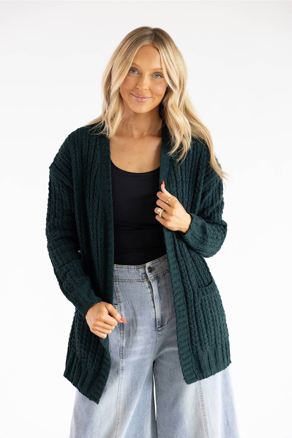 A woman modeling a dark green cable-knit open-front cardigan over a black top and light-wash jeans, photographed against a plain white studio background.