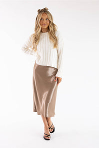A woman modeling a white ribbed knit long-sleeve top and a shiny taupe midi skirt, paired with black strappy heels, against a plain white studio background.