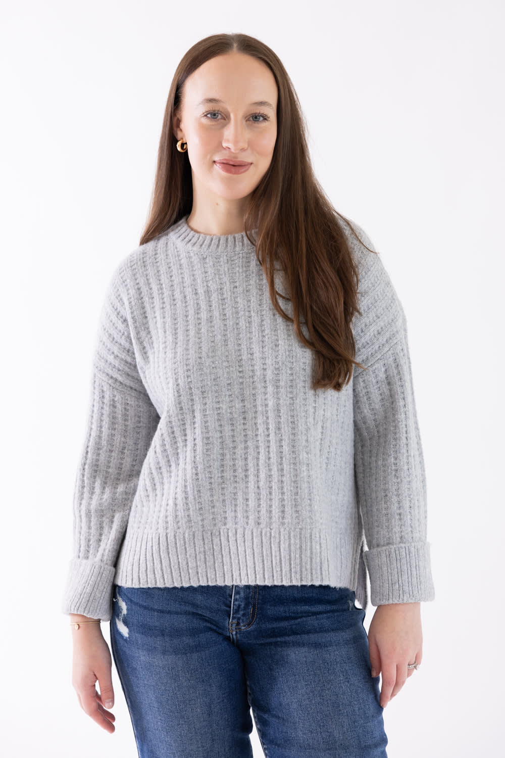 A woman wearing a light gray ribbed knit sweater and blue jeans, standing against a plain white studio background.