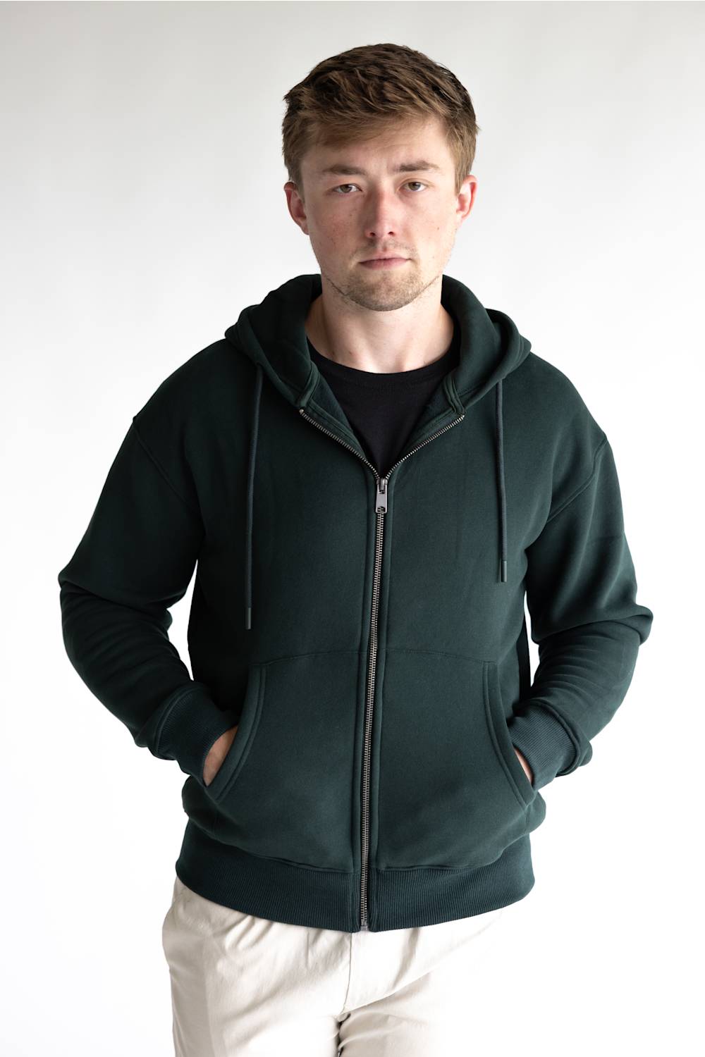 A man wearing a dark forest green zip-up hoodie over a black t-shirt and off-white trousers, standing against a plain light gray background.