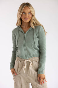 A woman modeling a sage green ribbed knit zip-up hoodie with a front pocket, paired with beige drawstring pants, against a plain white studio background.