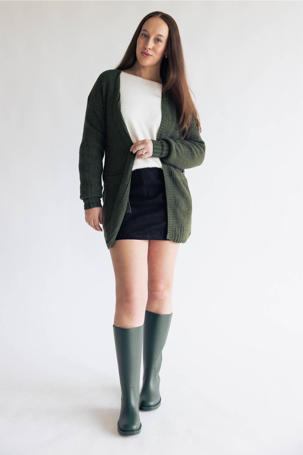 A woman modeling a dark green knit cardigan over a white top and black skirt, paired with dark green knee-high boots, against a plain white studio background.