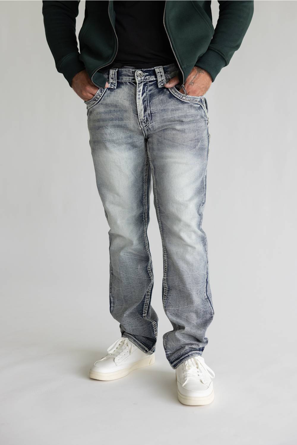 A person wearing light-wash, faded blue denim jeans with a straight-leg fit, styled with a dark green zip-up hoodie, black t-shirt, and white sneakers against a light gray studio background.