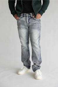 A person wearing light-wash, faded blue denim jeans with a straight-leg fit, styled with a dark green zip-up hoodie, black t-shirt, and white sneakers against a light gray studio background.