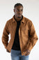A man wearing a brown button-up shirt jacket over a black t-shirt and blue jeans, photographed against a plain white studio background.