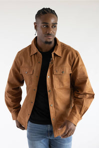 A man wearing a brown button-up shirt jacket over a black t-shirt and blue jeans, photographed against a plain white studio background.