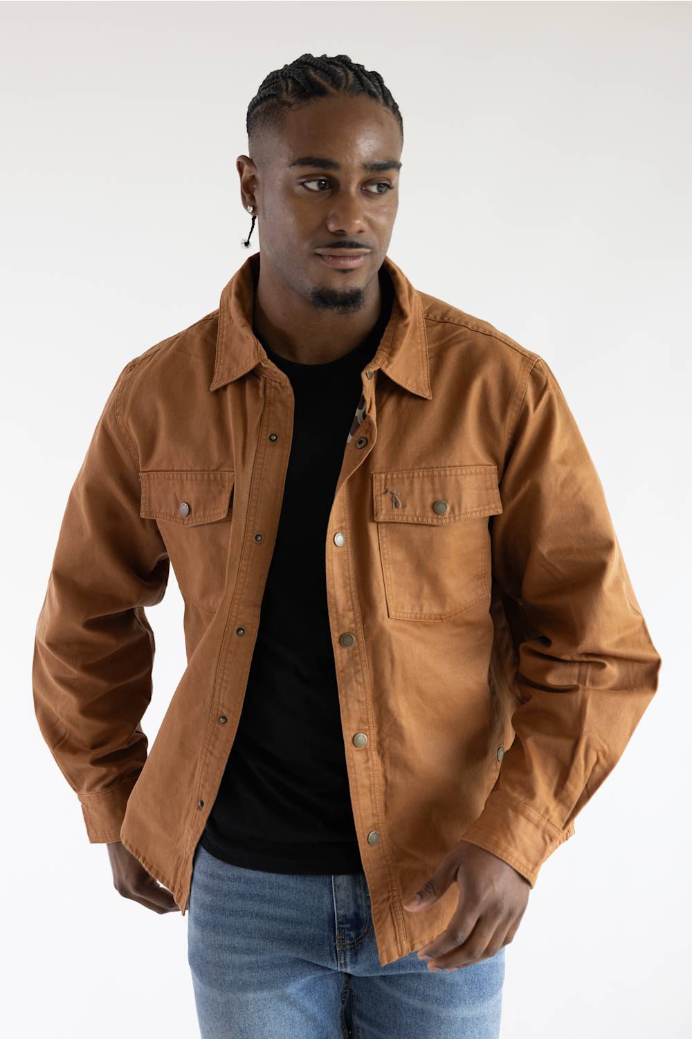 A man wearing a brown button-up shirt jacket over a black t-shirt and blue jeans, photographed against a plain white studio background.