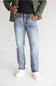 A person wearing light-wash blue jeans, a dark navy T-shirt, an olive green jacket, and white sneakers, standing against a plain light gray studio background.