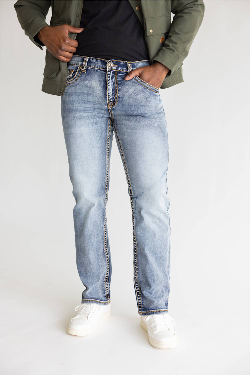A person wearing light-wash blue jeans, a dark navy T-shirt, an olive green jacket, and white sneakers, standing against a plain light gray studio background.