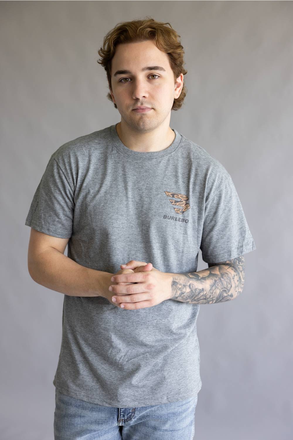 A man wearing a heather gray crewneck t-shirt with a small graphic on the left chest, paired with light blue jeans, standing against a neutral gray background.
