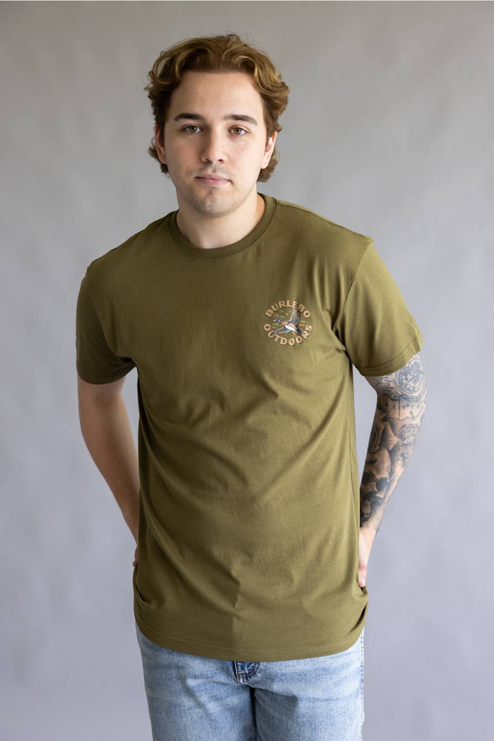 A man wearing an olive green crewneck t-shirt with a small circular graphic on the left chest, paired with light blue jeans, standing against a neutral gray studio background.