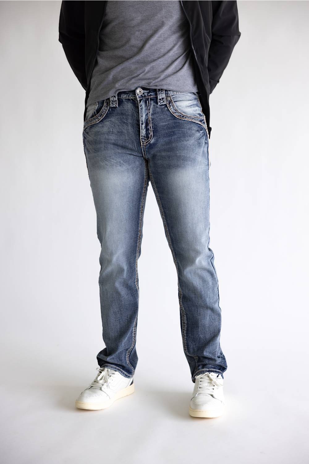 A person wearing light-to-medium wash blue jeans with a gray t-shirt, black jacket, and white sneakers, shown from the waist down against a plain light gray studio background.