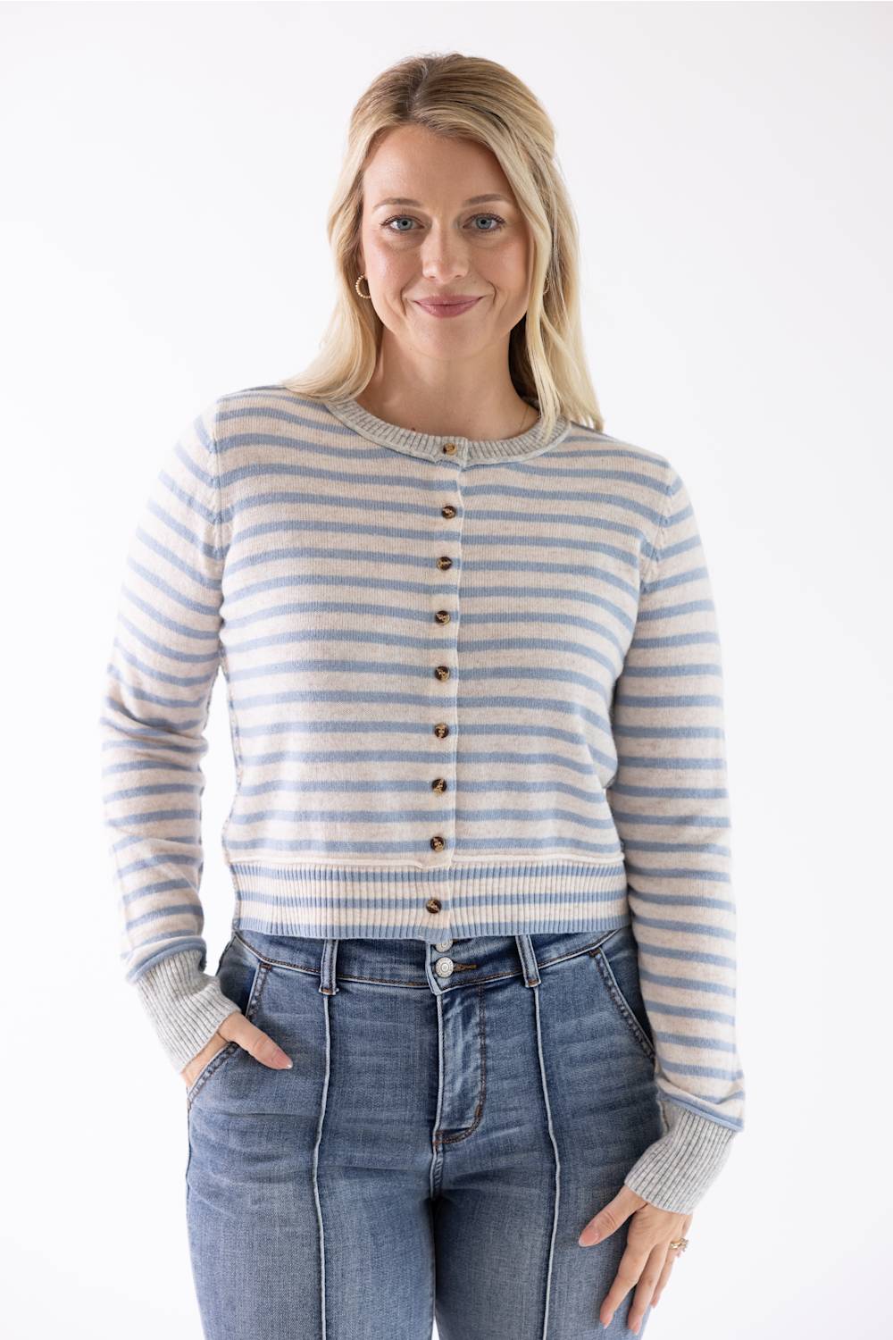A woman modeling a light blue and beige striped button-up cardigan sweater with blue jeans, photographed against a plain white studio background.