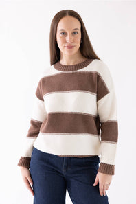 A woman wearing a brown and cream striped knit sweater with dark blue jeans, standing against a plain white studio background.