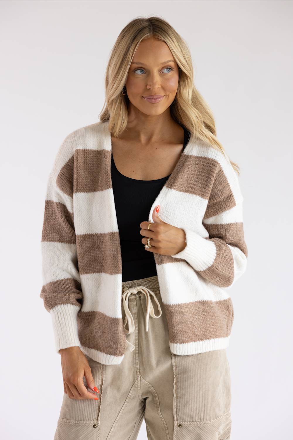 A blonde woman modeling a long-sleeve knit cardigan featuring bold white and tan horizontal stripes, layered over a black scoop-neck shirt and light beige drawstring pants.