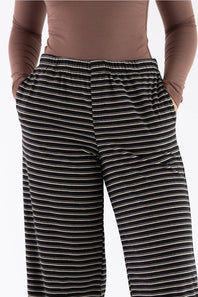 A close-up of a person wearing black wide-leg pants with thin white horizontal stripes, paired with a brown long-sleeve top, against a plain light gray background.
