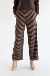 A person wearing brown wide-leg pants with thin beige horizontal stripes, paired with a dark brown sweater and beige heels, shown from the waist down against a plain white studio background.