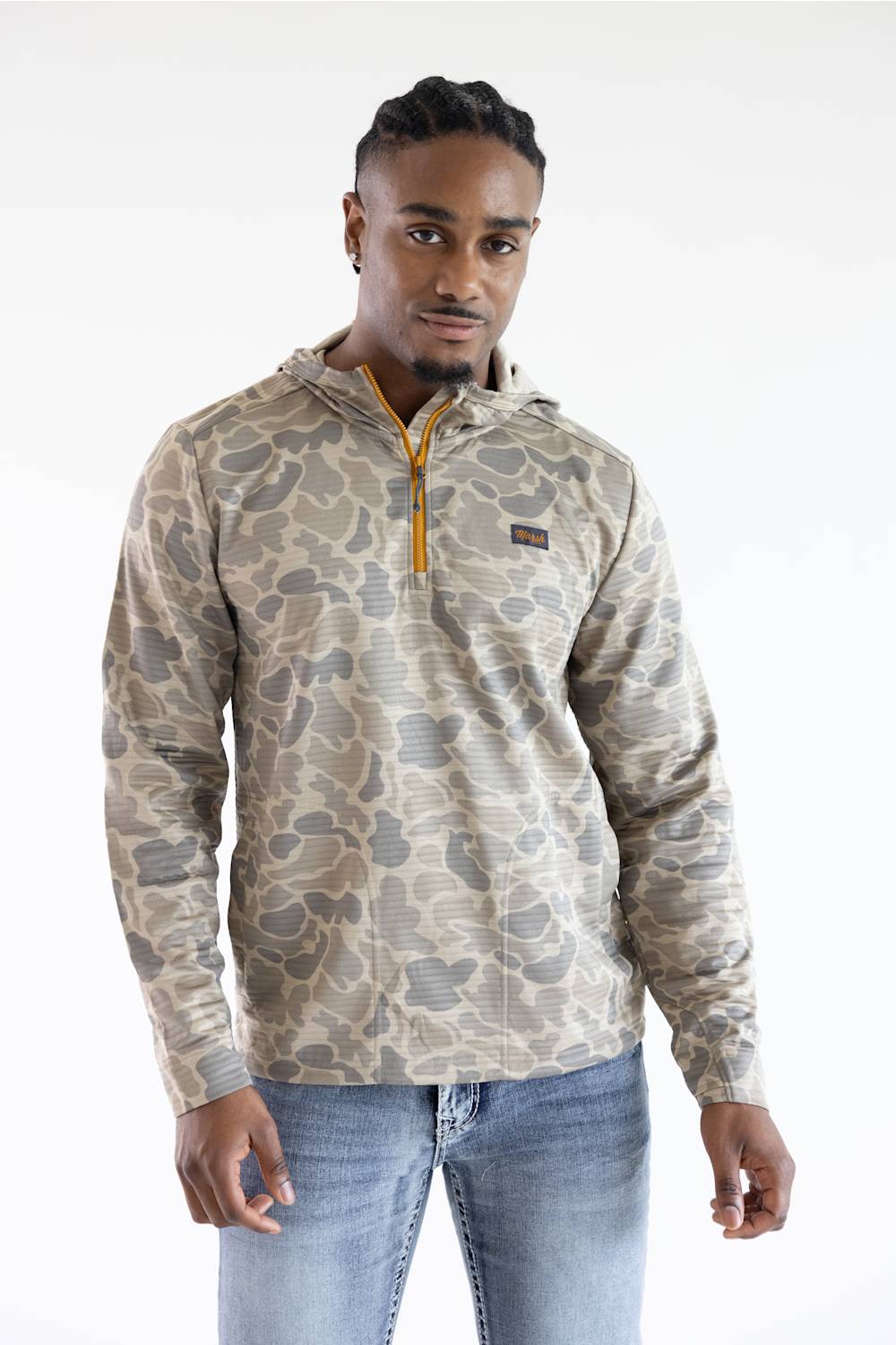 A man wearing a beige and gray camouflage quarter-zip pullover hoodie with a small orange zipper pull and a small chest logo patch, paired with blue jeans, against a plain white background.