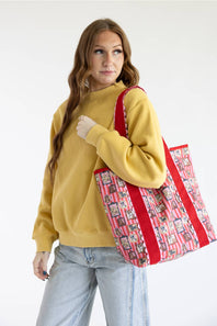 A woman wearing a mustard yellow sweatshirt and light blue jeans carries a red and pink patterned tote bag with red straps against a plain white studio background.