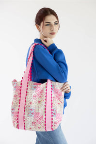 A woman wearing a blue sweatshirt and jeans carries a large tote bag with a pink and white floral pattern and pink straps, set against a plain white background.