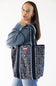 A woman wearing a blue sweatshirt and jeans carries a navy blue tote bag with a colorful floral pattern and black shoulder straps with white scalloped trim, against a plain white background.
