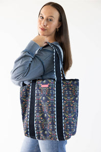 A woman wearing a blue sweatshirt and jeans carries a navy blue tote bag with a colorful floral pattern and black shoulder straps with white scalloped trim, against a plain white background.