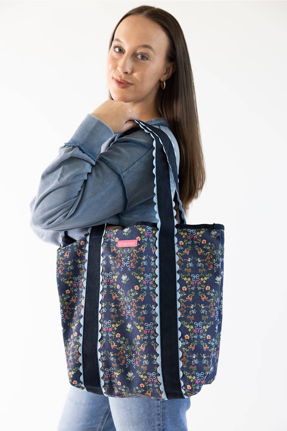 A woman wearing a blue sweatshirt and jeans carries a navy blue tote bag with a colorful floral pattern and black shoulder straps with white scalloped trim, against a plain white background.