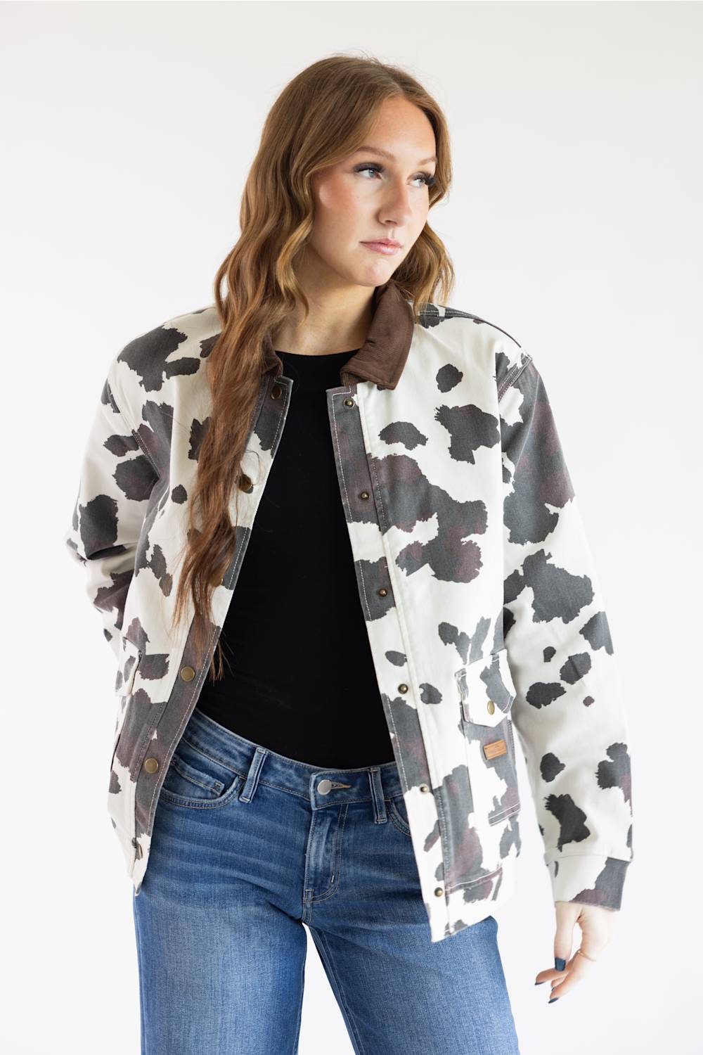 A woman modeling a cow-print jacket with a brown collar, worn over a black top and blue jeans, against a plain white studio background.