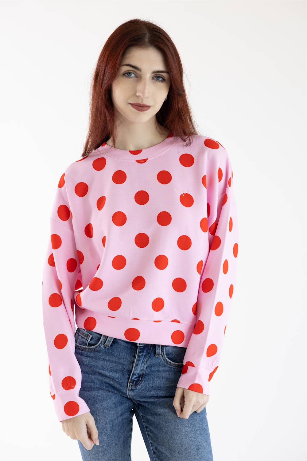 A woman with red hair wearing a pink long-sleeve sweatshirt with large red polka dots, paired with blue jeans, against a plain white background.