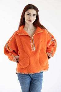 A woman wearing an oversized bright orange fleece pullover with floral patterned trim on the collar, zipper placket, cuffs, and hem, paired with blue jeans, against a plain white background.