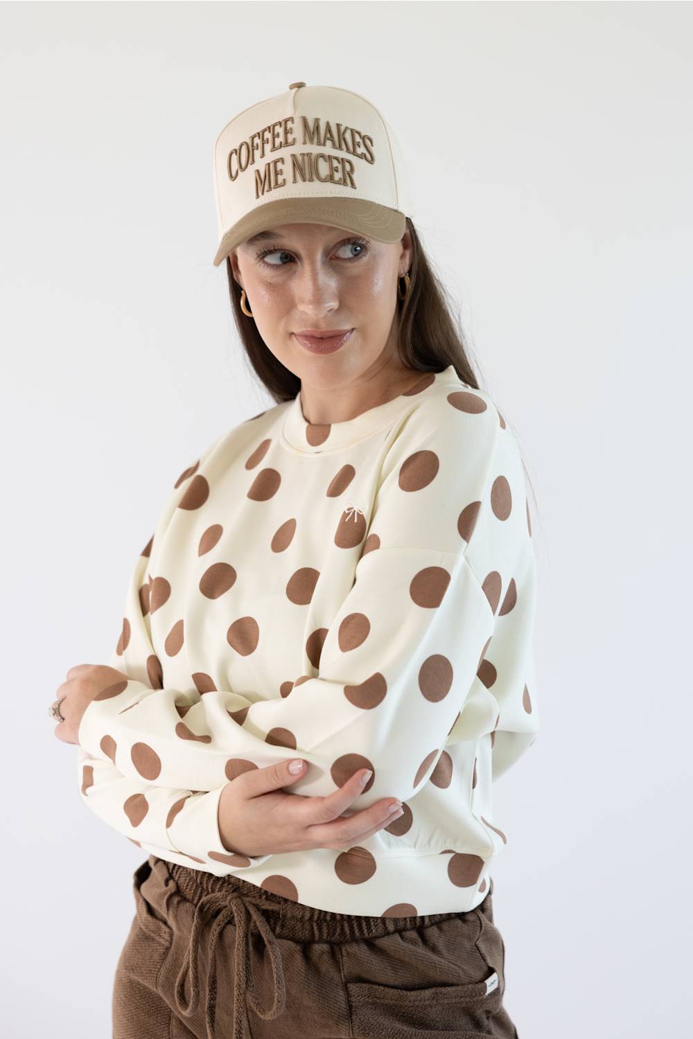 A woman wearing a cream sweatshirt with large brown polka dots, brown pants, and a beige baseball cap with the text 'COFFEE MAKES ME NICER' in brown, posed against a plain white studio background.